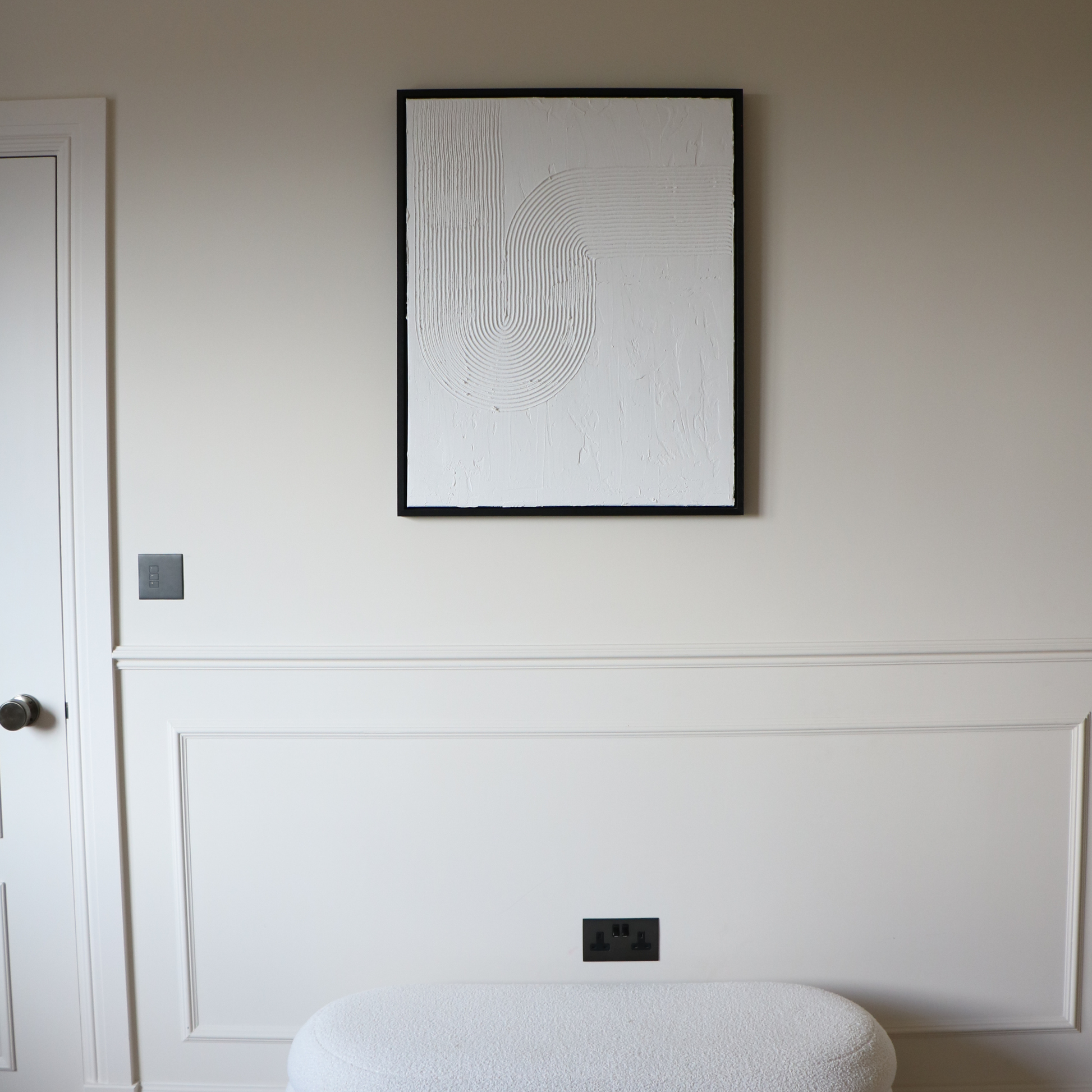 Framed abstract artwork on a wall with white paneling and a white ottoman.